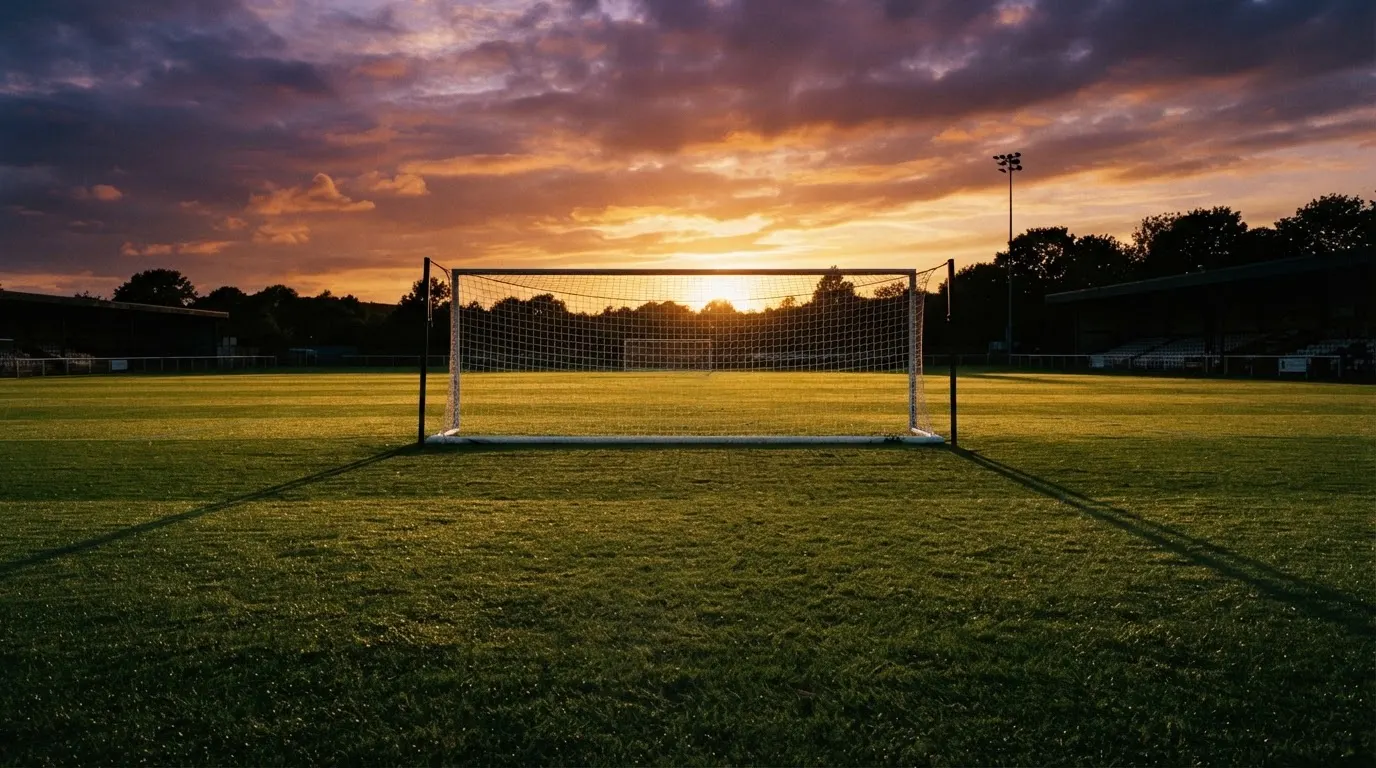 Ein Fußballtor mit Netz bei Sonnenuntergang auf einem leeren Platz, die Schatten der Torpfosten liegen lang auf dem Gras.