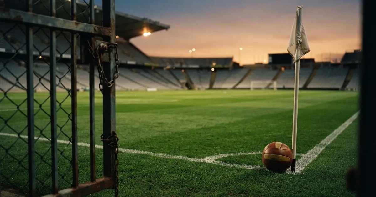 Blick durch ein Stadiontor auf einen grünen Rasen im Abendlicht, ein Ball ruht an der Eckfahne, die Tribünen sind leer.