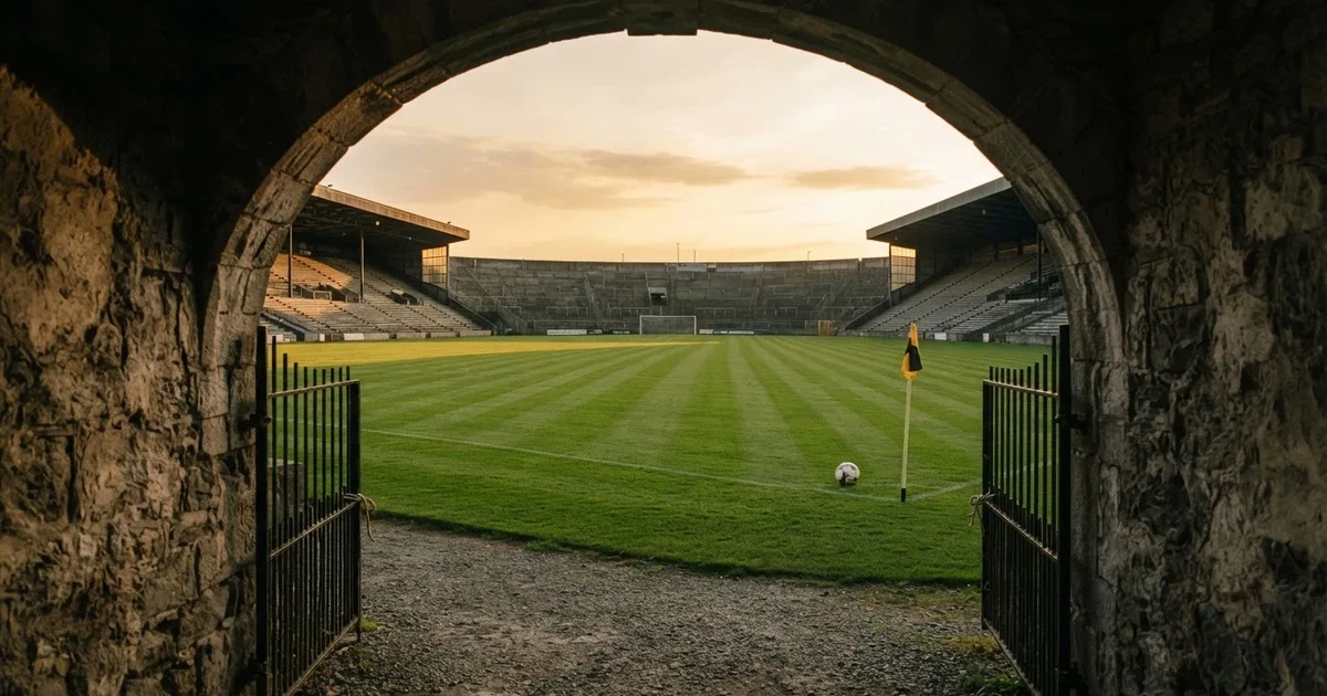 Blick durch ein Stadiontor auf einen grünen Rasen im Abendlicht, ein Ball liegt an der Eckfahne, die Ränge sind leer.