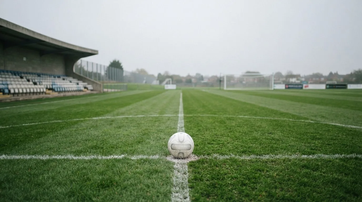 Ein Fußball liegt auf dem Anstoßpunkt eines leeren Rasenplatzes, im Hintergrund eine ruhige Tribüne unter bedecktem Himmel.