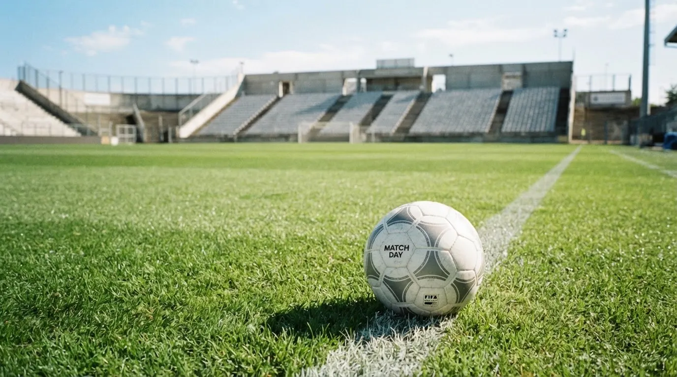 Ein Fußball liegt am Spielfeldrand eines Rasenplatzes, im Hintergrund eine leere Tribüne unter hellem Himmel.