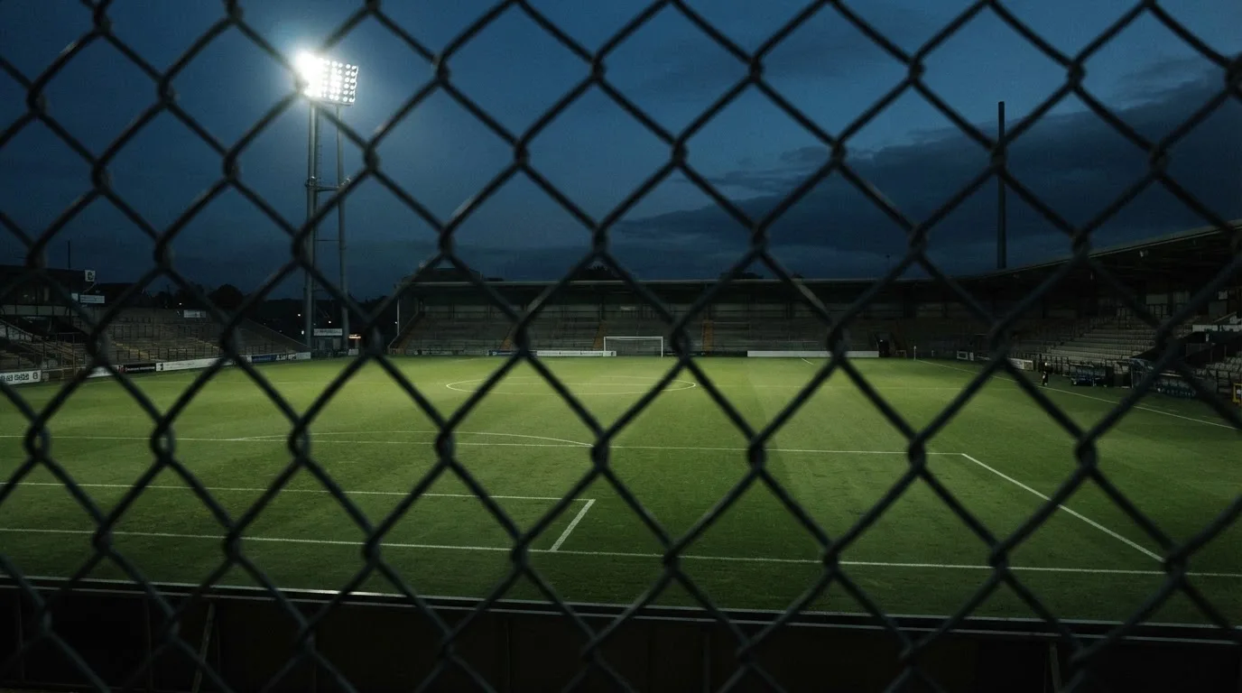 Eine Flutlichtanlage über einem leeren Sportplatz am Abend, im Vordergrund ein Maschendrahtzaun.