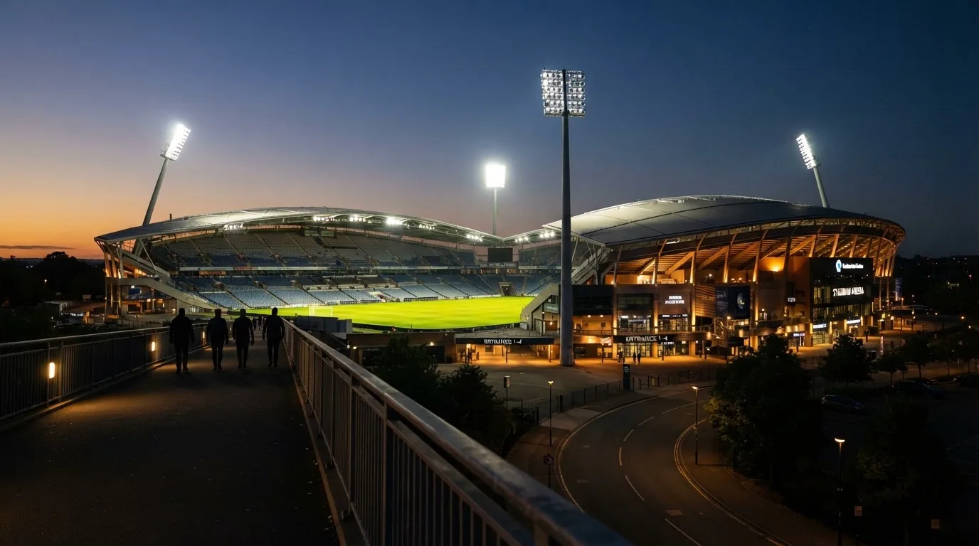Ein Stadion bei Dämmerung mit eingeschalteten Flutlichtmasten, von einer Brücke aus aufgenommen.