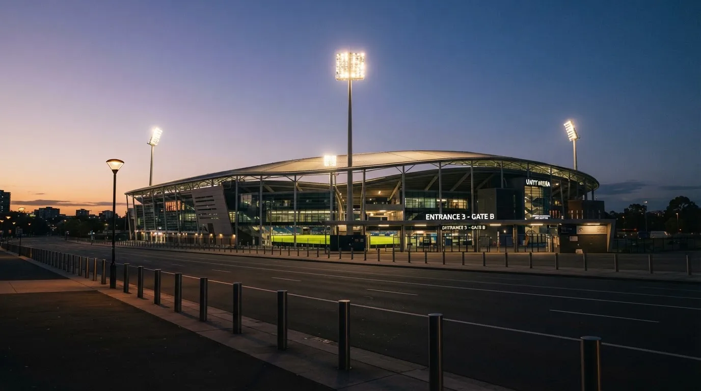 Ein Stadion aus der Ferne bei Flutlicht, davor eine ruhige Straße in der Abenddämmerung.