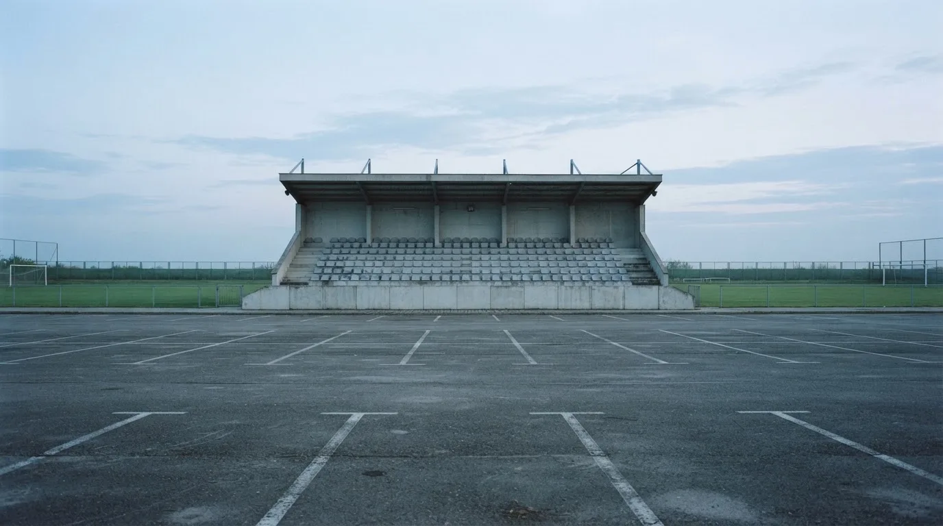 Eine leere Tribüne eines kleinen Stadions am frühen Morgen, aus der Ferne über einen menschenleeren Parkplatz fotografiert.