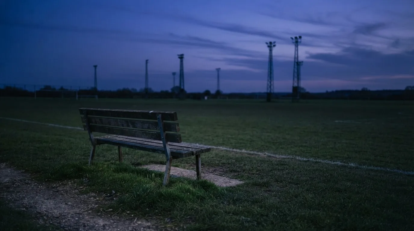 Eine leere Parkbank am Rand eines Fußballfelds in der Abenddämmerung, im Hintergrund ragen dunkle Flutlichtmasten in den Himmel.