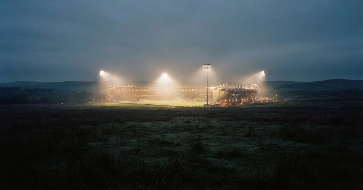 Ein Stadion unter Flutlicht, aus großer Entfernung über eine dunkle Wiese fotografiert, am Himmel zieht Nebel auf.