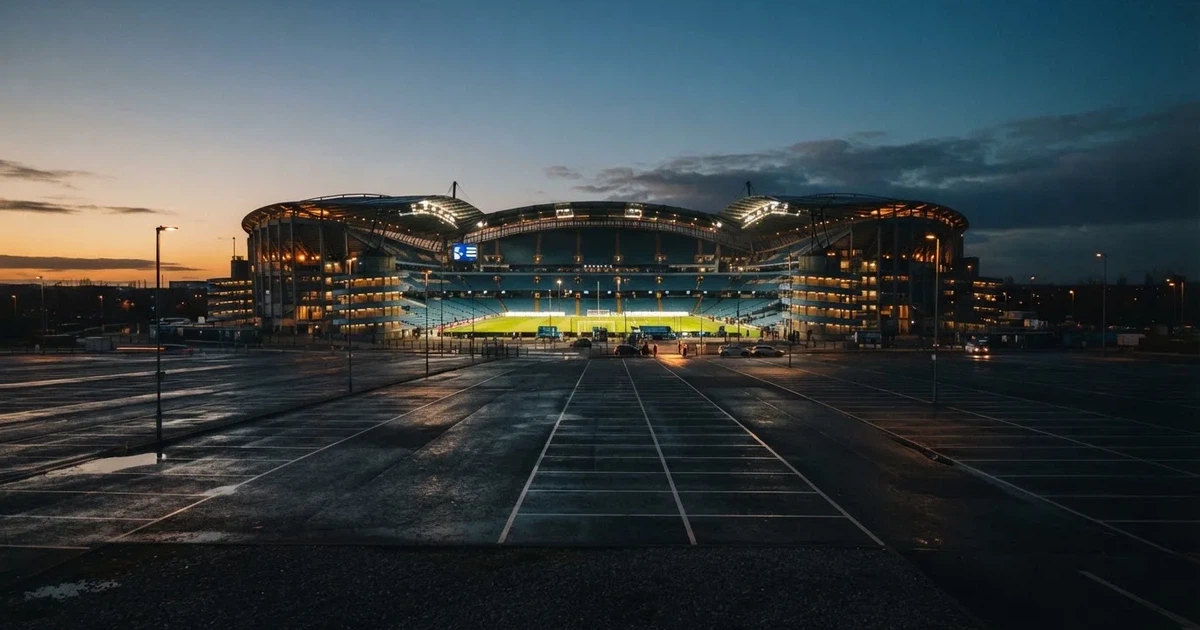 Ein Fußballstadion unter Flutlicht aus großer Entfernung, davor ein dunkler Parkplatz und ein heller Abendhimmel.