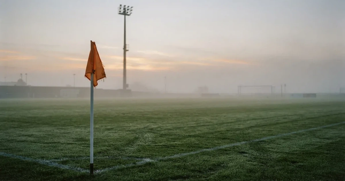 Eine Eckfahne steht im Nebel am Rand eines leeren Rasenplatzes in der Morgendämmerung, dahinter ein Flutlichtmast.