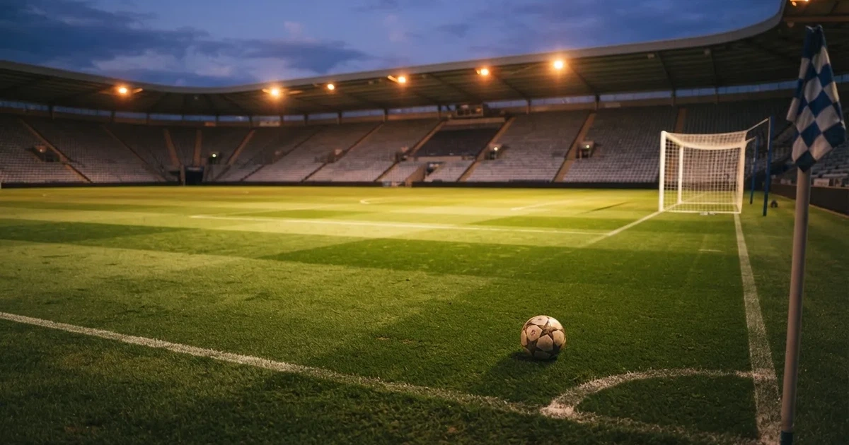 Ein Fußballplatz mit frisch gemähtem Rasen im Abendlicht, ein Ball liegt im Strafraum, im Hintergrund unscharf eine leere Tribüne.