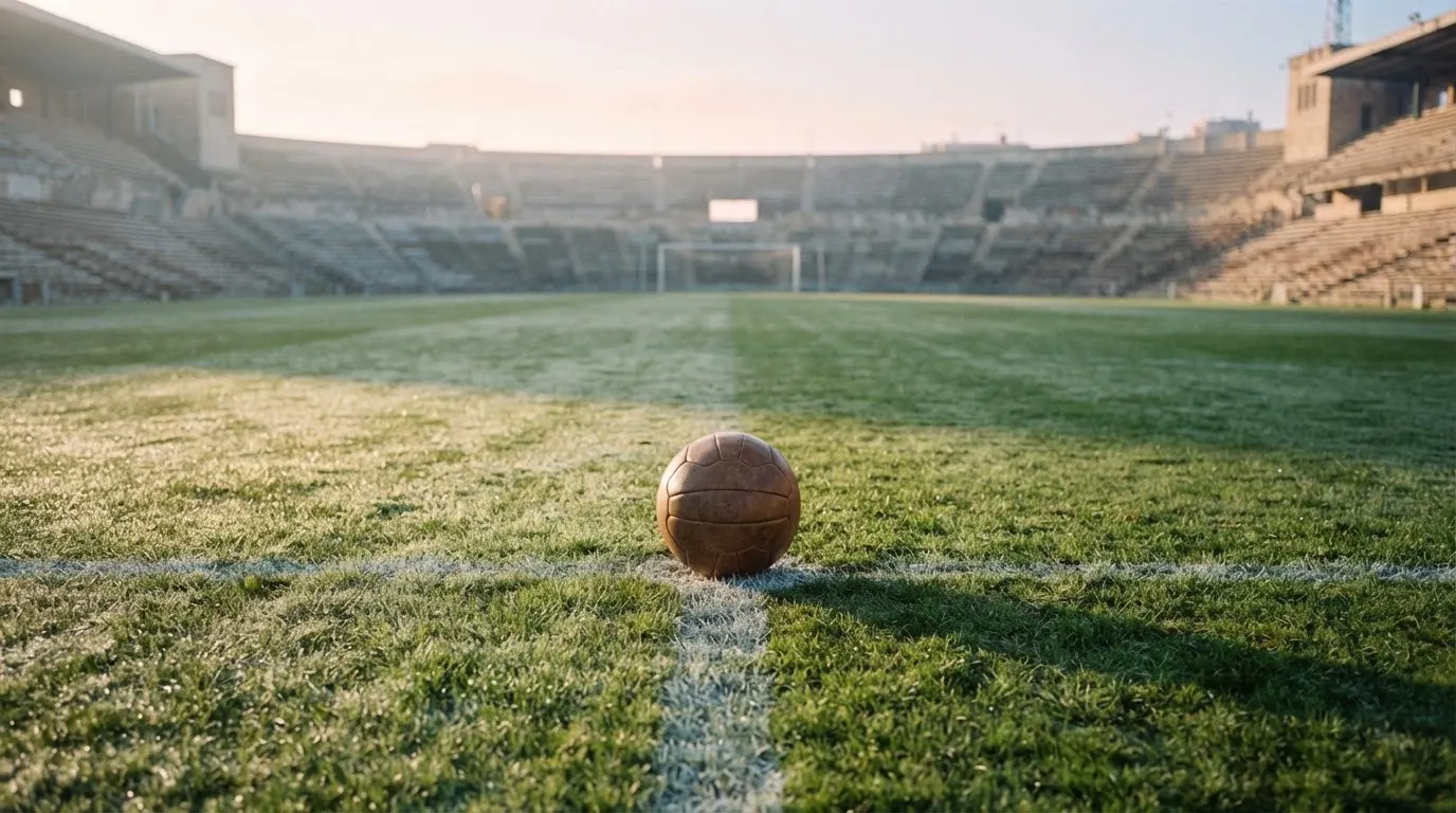 Ein Fußball liegt auf dem Anstoßpunkt eines leeren Rasenplatzes im frühen Morgenlicht, dahinter eine leere Tribüne.