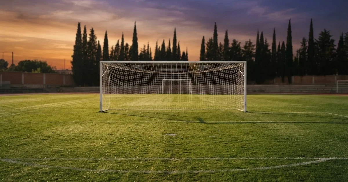 Ein Fußballtor mit Netz auf einem leeren Platz im Abendlicht, der Rasen frisch gemäht, im Hintergrund Zypressen.