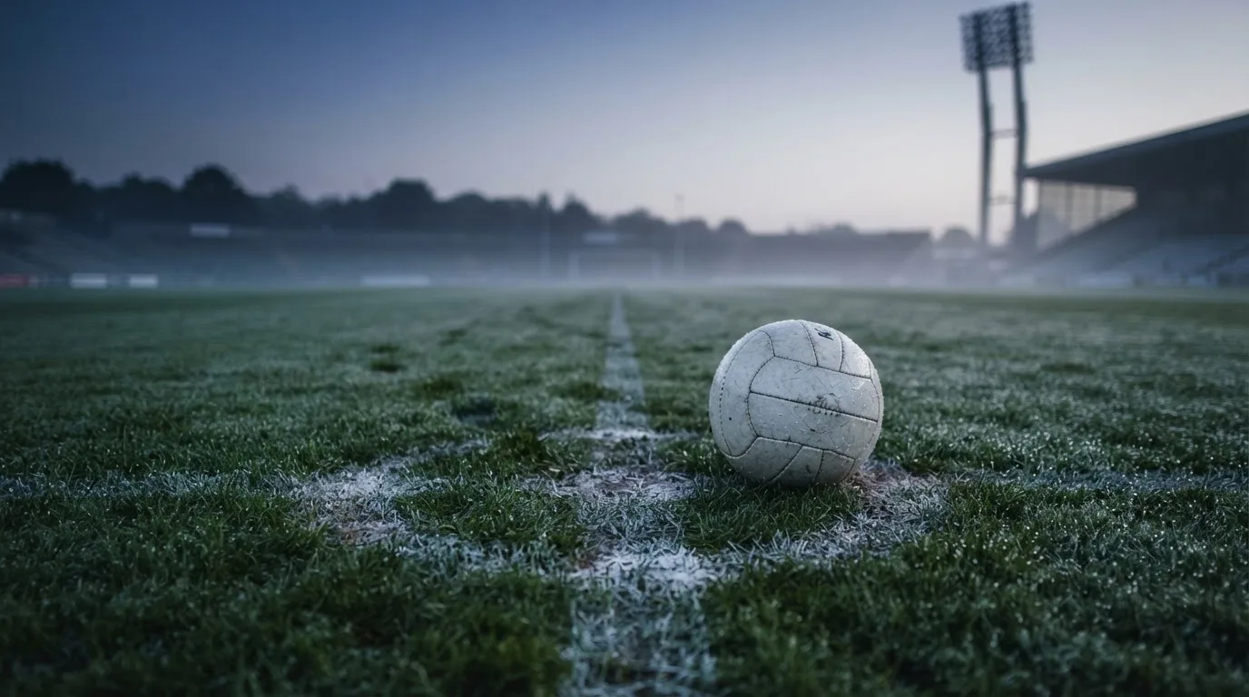 Ein Fußball ruht auf dem Mittelkreis eines leeren Rasenplatzes in der Morgendämmerung, Tau liegt auf dem Gras.