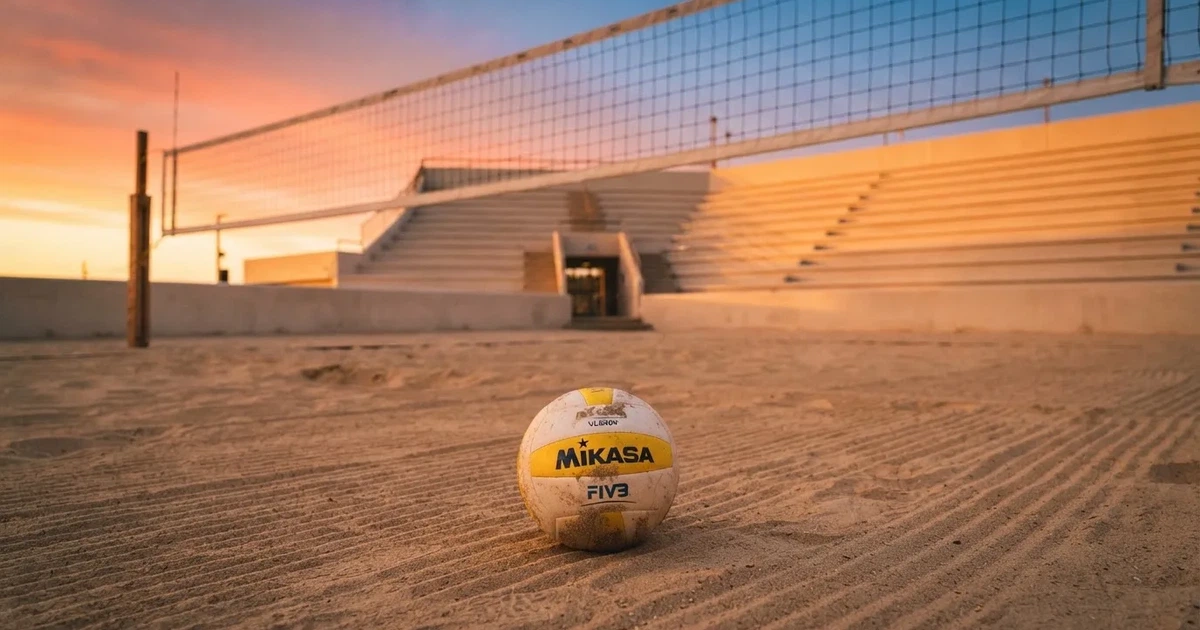 Ein Volleyball liegt auf dem Sand eines Beachvolleyball-Feldes neben dem gespannten Netz, im Hintergrund eine leere Tribüne bei Abendlicht.