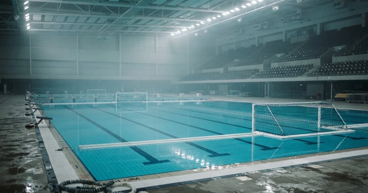 Ein Schwimmbecken mit türkisfarbenem Wasser und zwei schwimmenden Torrahmen am gegenüberliegenden Beckenrand, menschenleer unter Hallenlicht.