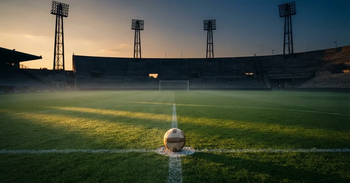 Ein Fußball liegt auf dem Anstoßpunkt eines leeren Rasenplatzes, im Hintergrund eine Tribüne und Flutlichtmasten in der Abenddämmerung.