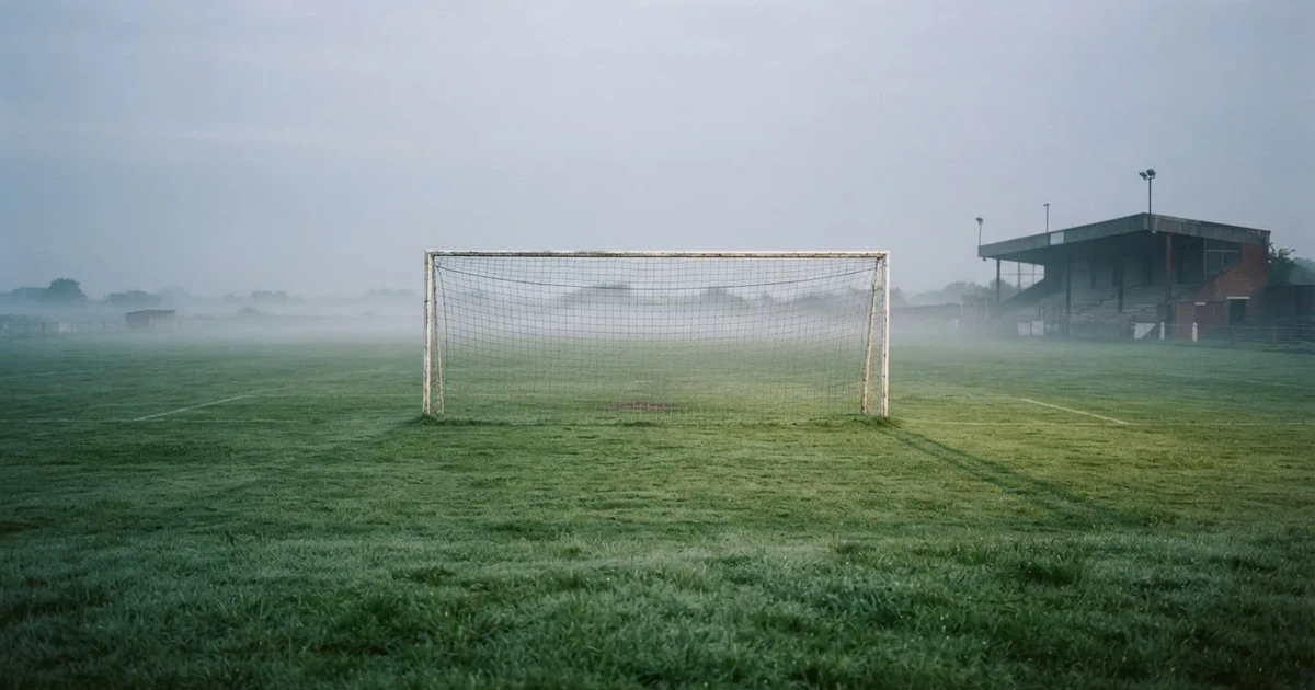 Ein Fußballtor mit Netz steht im Morgennebel auf einem leeren Rasenplatz, im Hintergrund die Silhouette einer kleinen Tribüne.