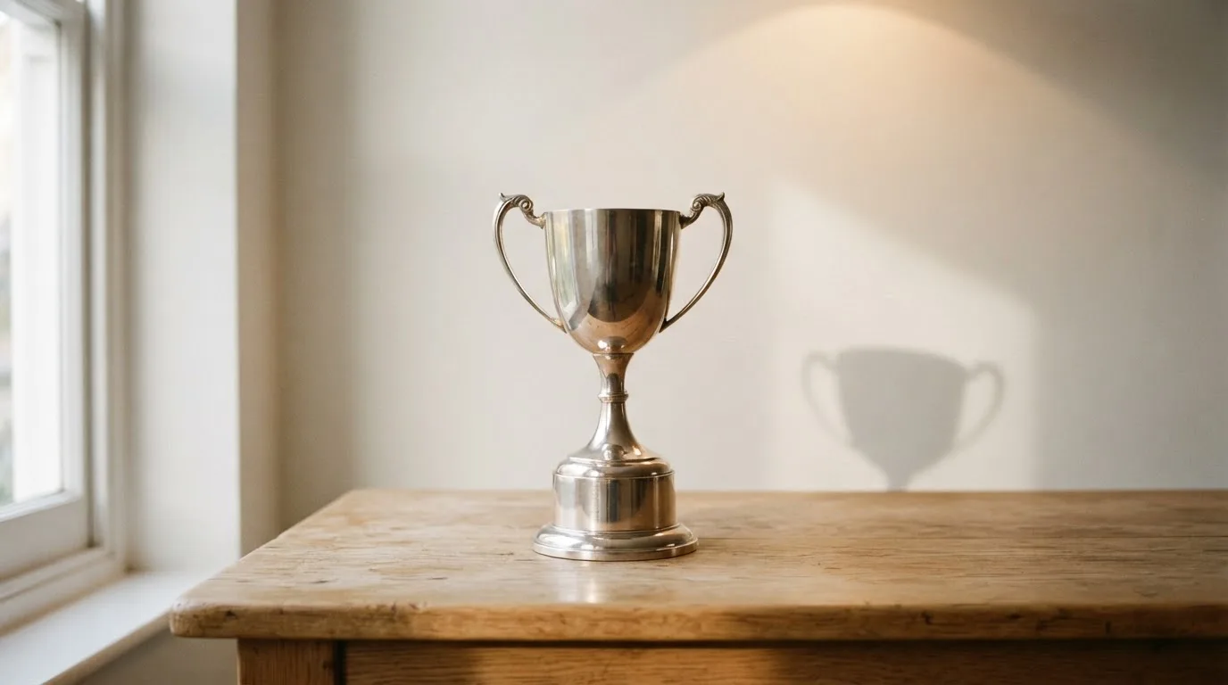 Ein Pokal ohne Gravur steht allein auf einem Holztisch vor einer hellen Wand, davon fällt ein weicher Schatten.
