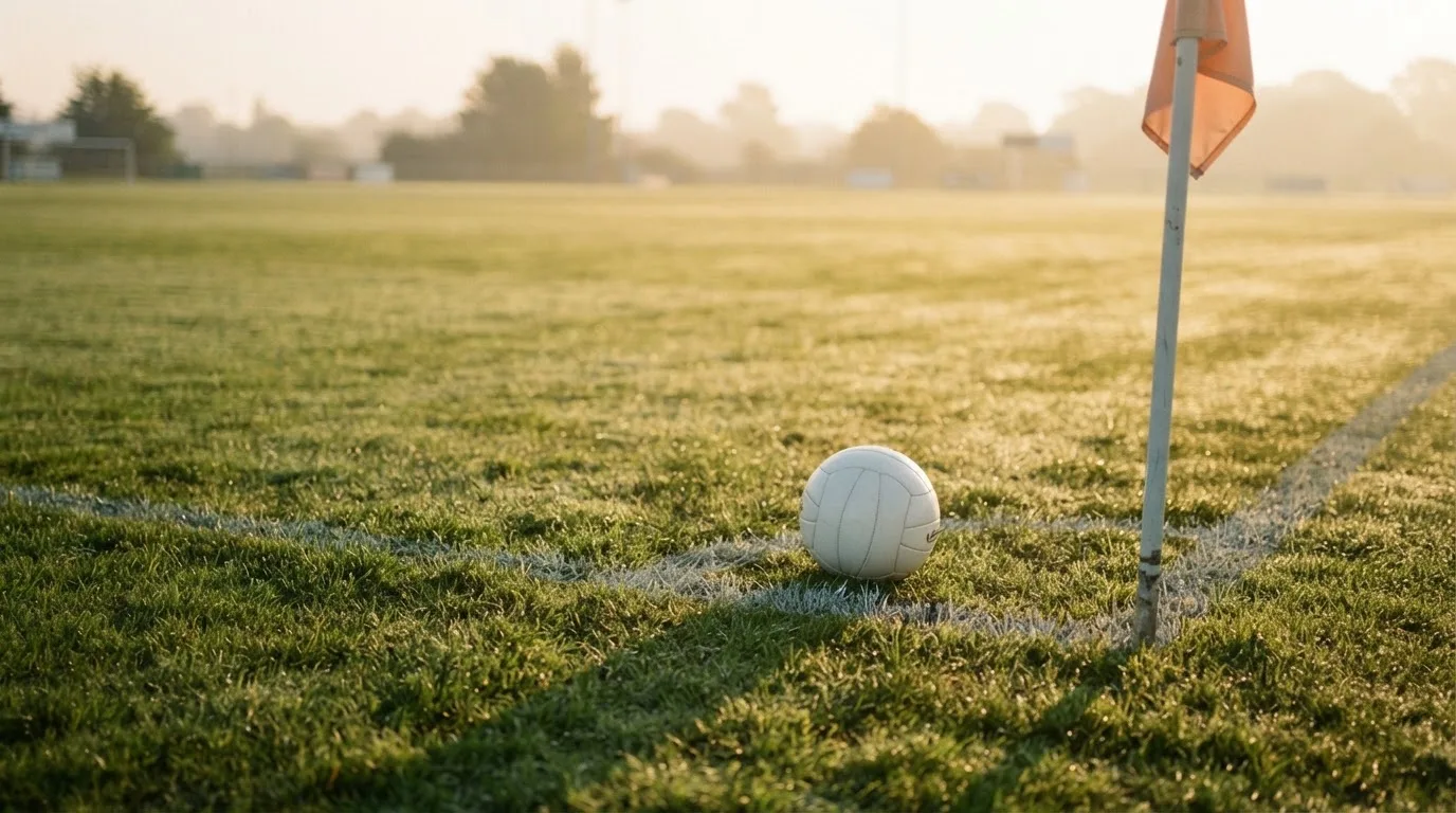 Ein Fußball liegt auf Rasen neben einer Eckfahne ohne Aufdruck auf einem leeren Platz im Morgenlicht.