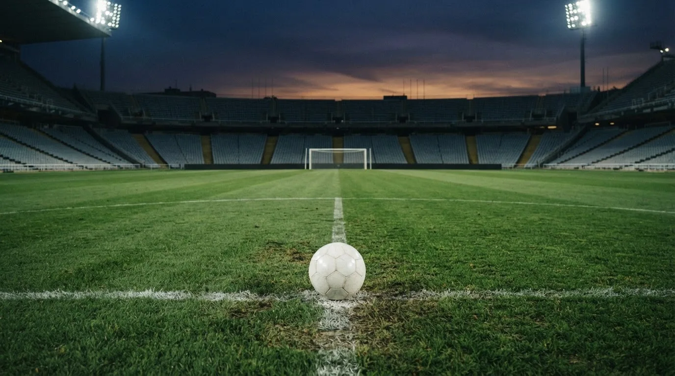 Ein Fußball liegt auf dem Anstoßpunkt eines Rasenplatzes, dahinter eine leere Tribüne im Abendlicht.