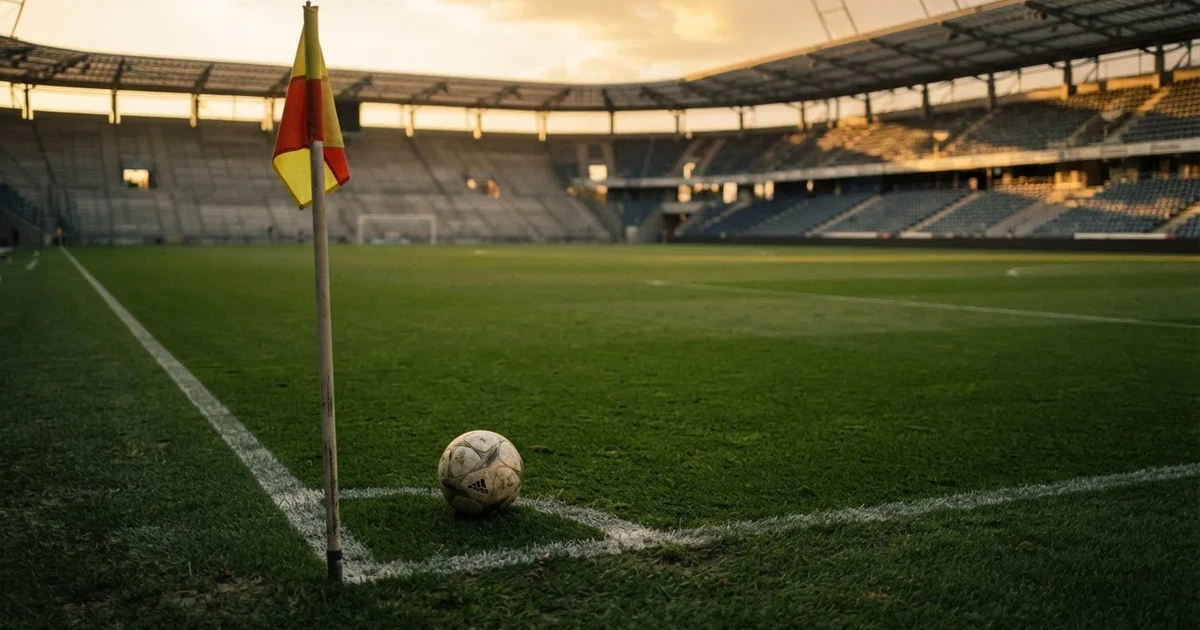 Ein Fußball liegt am Viertelkreis an der Eckfahne eines Rasenplatzes, im Hintergrund leere Tribünen in warmem Abendlicht.