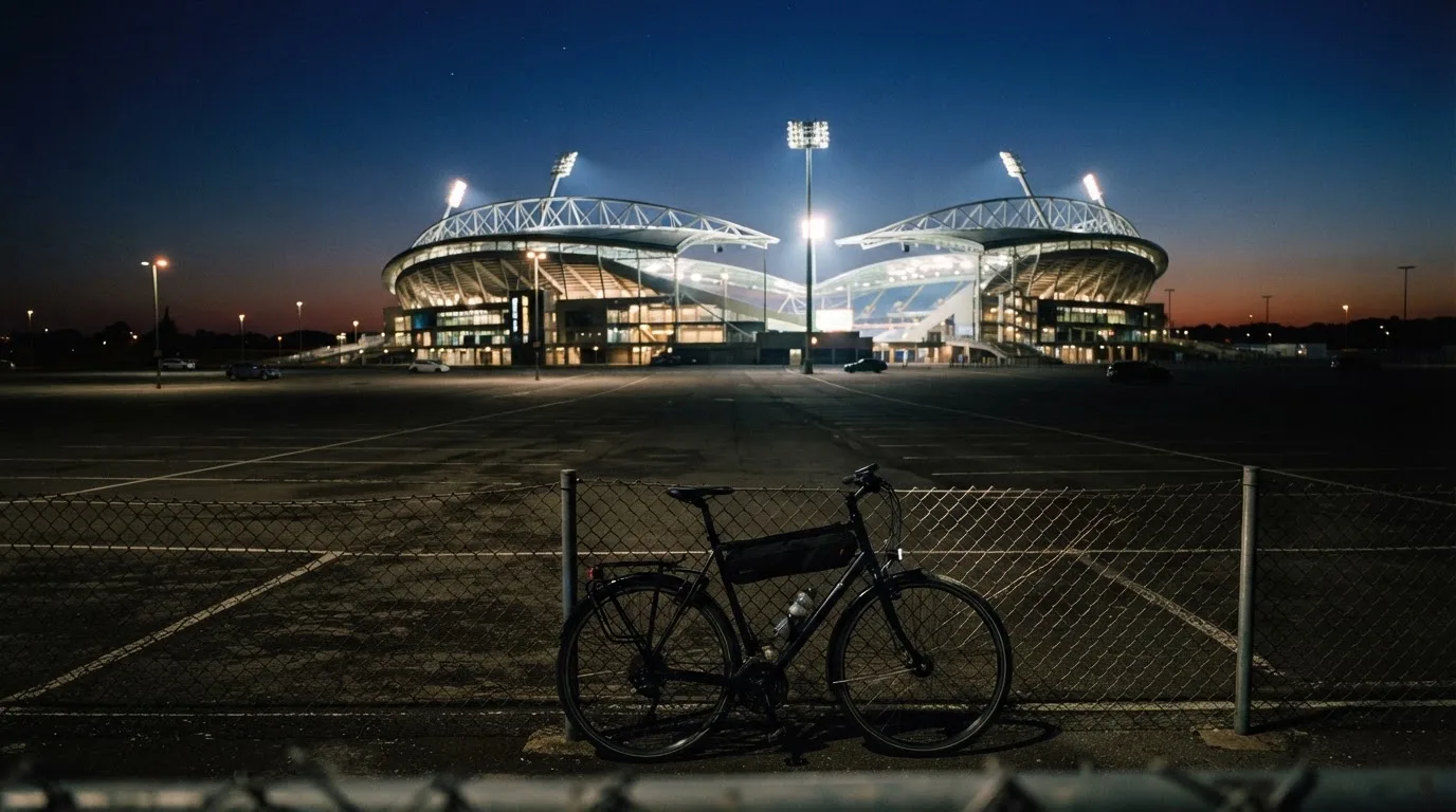 Ein Stadion unter Flutlicht aus der Ferne gesehen, davor ein dunkler Parkplatz und ein einzelnes Fahrrad an einem Zaun.