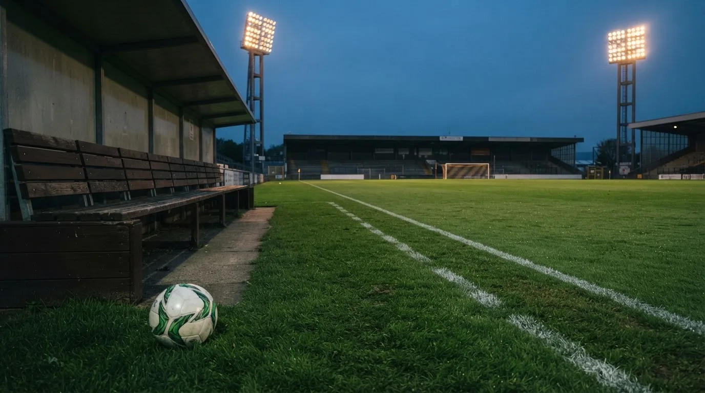 Ein Fußball liegt am Spielfeldrand neben einer leeren Ersatzbank, das Flutlicht über dem Platz leuchtet in der Dämmerung.